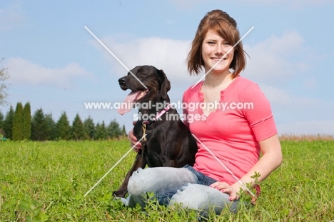 Young woman sitting with her black Labrador Retriever in a grassy field.