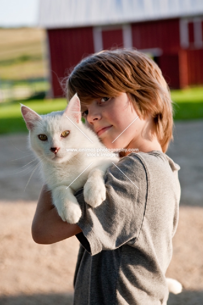Young boy holding a white cat near a red barn.