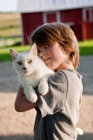 Picture of Young boy holding a white cat near a red barn.