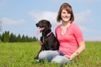 Picture of Young woman sitting with her black Labrador Retriever in a grassy field.